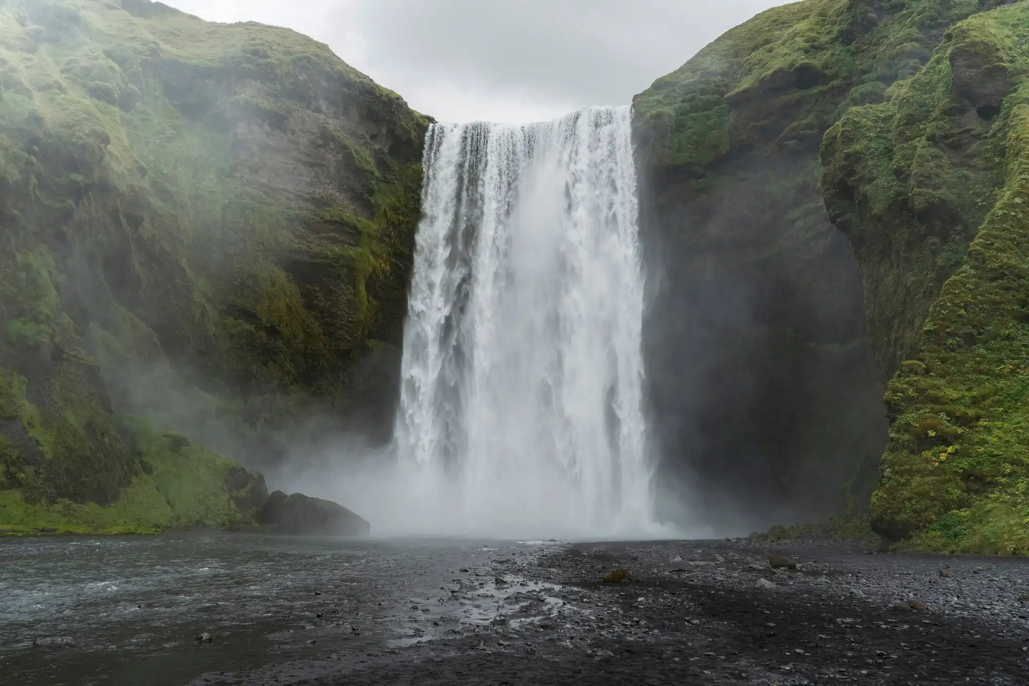 A wide view of a powerful waterfall plunging into a rocky riverbed, surrounded by steep green mountains and low mist.