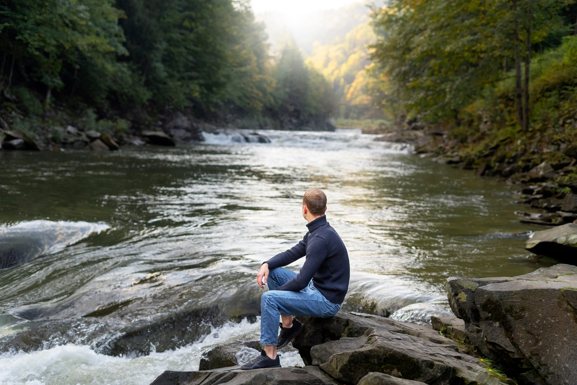 A man in a blue sweater and jeans sits on a rocky riverbank, looking out over a fast-flowing stream surrounded by dense green trees as sunlight filters through the canopy.